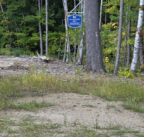 An image of a white sign with "Hassunnek Hill" in blue capital letters, framed by tall trees in the background and a patch of dirt and grass in the foreground.